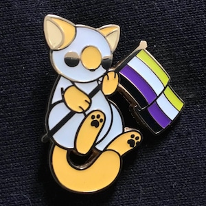 Peut inclure: Épingle en émail représentant un chat de dessin animé tenant un drapeau de la fierté non binaire. Le chat est blanc et jaune avec des détails noirs. Le drapeau a des rayures noires, violettes, blanches et jaunes. L'épingle a une bordure dorée.