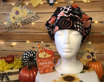 May include: A black and orange patterned bouffant scrub cap with pumpkin and plaid designs. The cap is displayed on a white mannequin head. The background includes a wooden wall, decorative pumpkins, and a sign that says "Happy Pumpkin Spice Season."