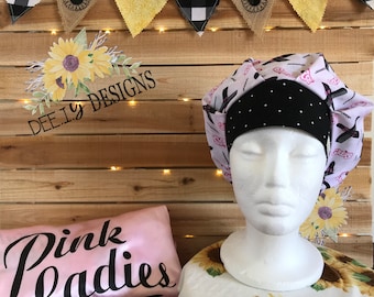 May include: A white mannequin head is wearing a bouffant surgical scrub cap with a black polka dot band. The cap has a light purple background with black witch-themed graphics. A pink satin pillow with the words "Pink Ladies" is visible.