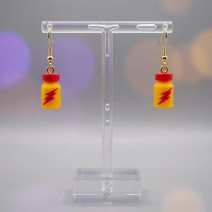 May include: A pair of yellow and red earrings shaped like small bottles, each with a red lightning bolt design. The earrings hang from gold-colored hooks and are displayed on a clear stand.