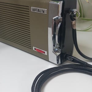 May include: An olive green and black Optalix device with a silver control panel and a red indicator light. A black USB cable with a USB-A connector is in the foreground. A plant in a blue pot is in the background.