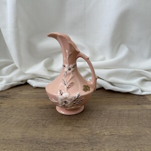 May include: A pink ceramic pitcher with a floral design. The pitcher has a wide mouth and a handle. The flowers are white with brown centres.