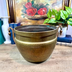May include: A vintage brass planter with two ornate handles. The planter has a hammered texture and a rounded shape. The brass has a warm, aged patina. The background includes a painting of roses and a small potted plant.