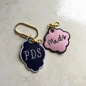 May include: Two cloud-shaped embroidered keychains with gold-tone hardware. One is dark navy blue with white stitching and the letters PDS. The other is light pink with black stitching and the name Mads written in a cursive script font.