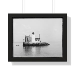 May include: A black and white photograph of a lighthouse situated on a small island. The lighthouse is white with a black roof and a black lantern. There are two smaller buildings on the island, one of which has a chimney. The island is surrounded by water.
