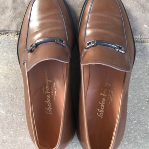 May include: A pair of brown leather loafers with a silver buckle detail. The loafers are made by Salvatore Ferragamo and have the words "Salvatore Ferragamo Studio" printed on the inside.