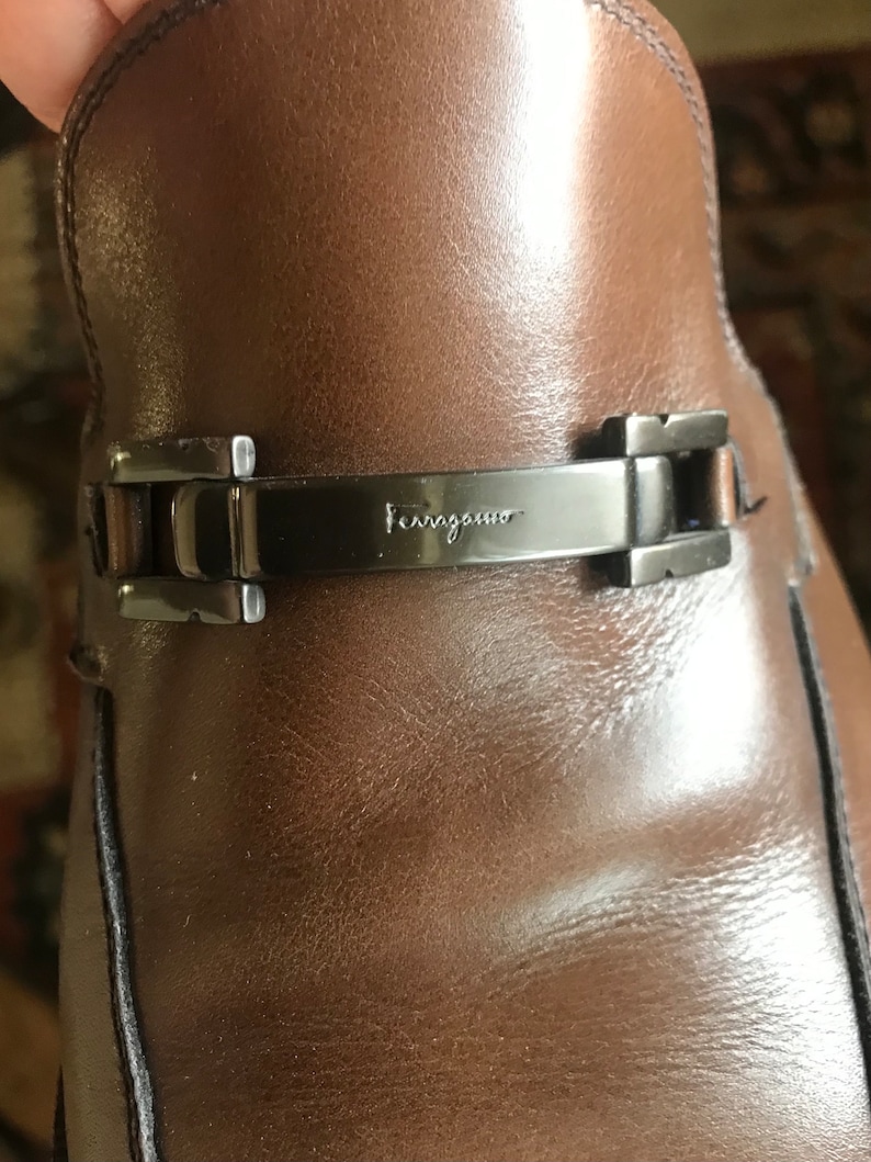 May include: Close-up of a brown leather shoe with a silver buckle and the word "Ferragamo" engraved on it.