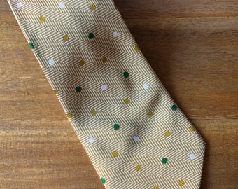 May include: A light yellow tie featuring a herringbone weave and small polka dots in white, green, and gold. The tie is made of a smooth fabric and is angled on a wooden surface.