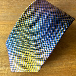 Peut inclure: Une cravate pliée avec un motif quadrillé. La cravate présente une palette de couleurs jaune, bleu, noir et violet. La cravate est faite d'un matériau brillant et est inclinée sur une surface en bois.
