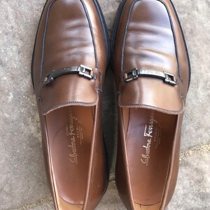 May include: A pair of brown leather loafers with a silver buckle detail. The loafers have a Salvatore Ferragamo STUDIO label on the inside.