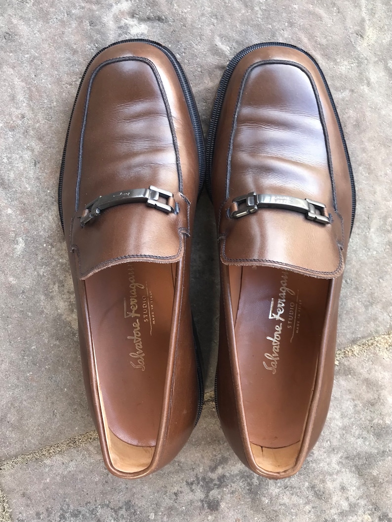May include: A pair of brown leather loafers with a silver buckle detail. The loafers have a Salvatore Ferragamo STUDIO label on the inside.