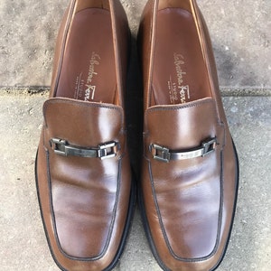 May include: A pair of brown leather loafers with silver buckles and the text "Salvatore Ferragamo Studio" on the inside of each shoe.