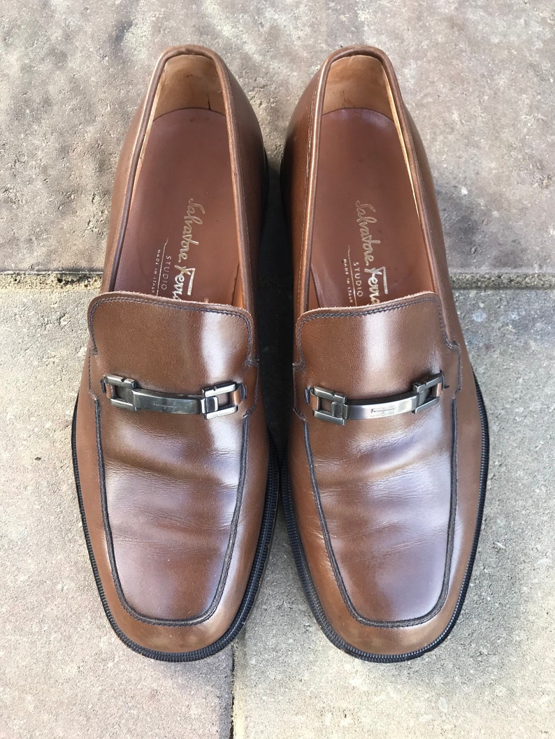 May include: A pair of brown leather loafers with silver buckles and the text "Salvatore Ferragamo Studio" on the inside of each shoe.