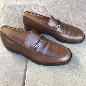 May include: A pair of brown leather loafers with a silver buckle detail. The loafers have a rounded toe and a low heel.