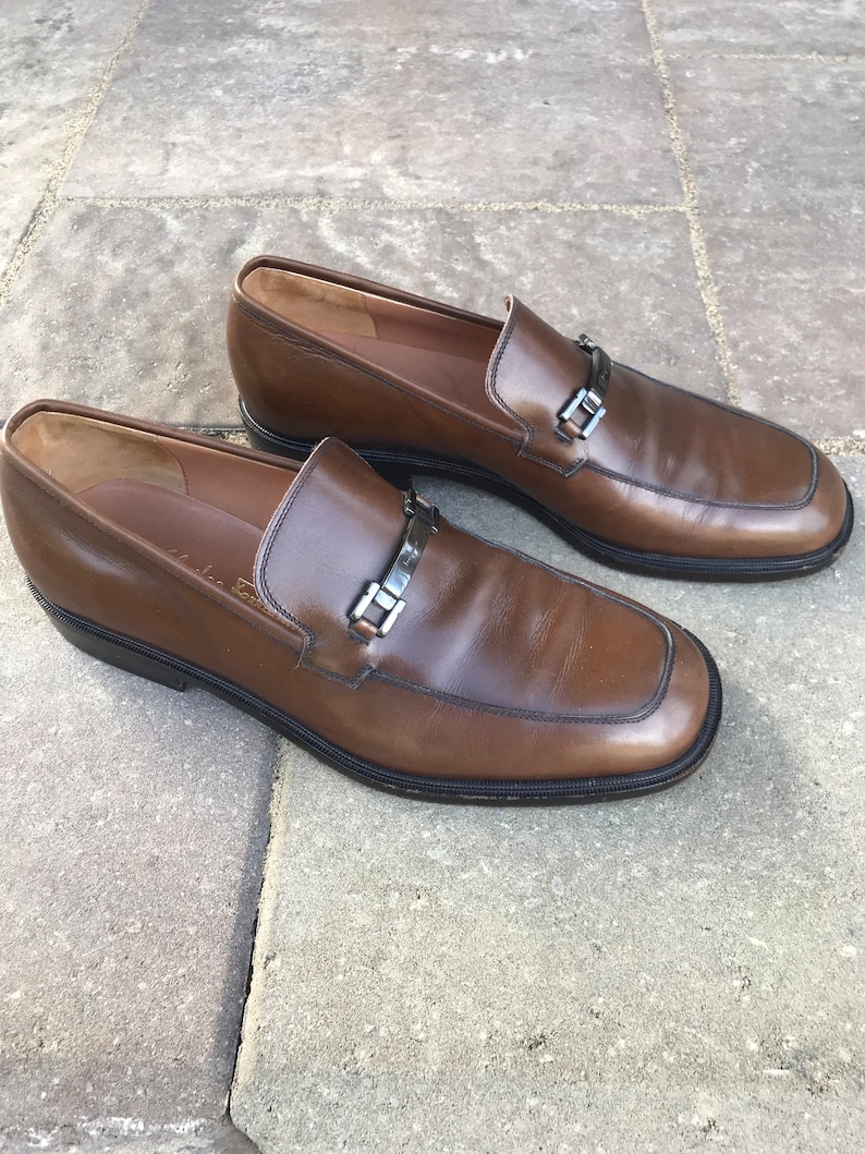 May include: A pair of brown leather loafers with a silver buckle detail. The loafers have a rounded toe and a low heel.