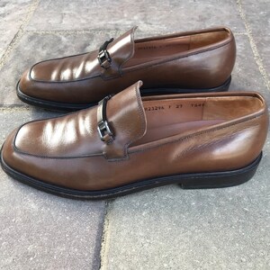 May include: A pair of brown leather loafers with a silver buckle detail. The loafers have a rounded toe and a low heel. The size is marked as 27.