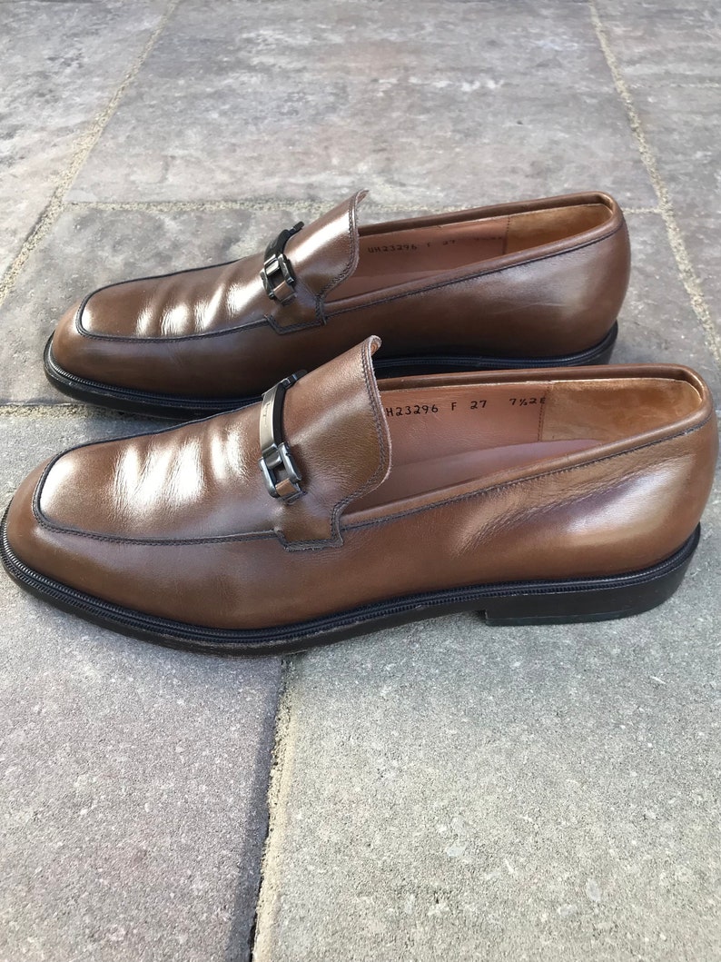 May include: A pair of brown leather loafers with a silver buckle detail. The loafers have a rounded toe and a low heel. The size is marked as 27.