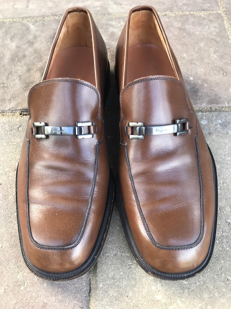 May include: A pair of brown leather loafers with a silver buckle detail. The loafers have a black rubber sole and a pointed toe.