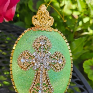 May include: A green Easter egg with a gold cross and rhinestones. The egg is decorated with glitter and has a gold stand.