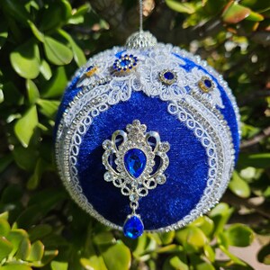 May include: A blue velvet Christmas ornament with white lace trim and silver and blue embellishments. The ornament features a crown-shaped design with a large blue jewel in the center.