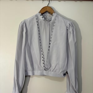May include: A light grey long-sleeve blouse featuring a high neckline, gathered shoulders, and a V-shaped decorative trim down the front. The garment has buttoned cuffs and a fitted waistband, presented on a wooden hanger against a wall.