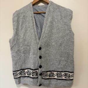 May include: A grey sleeveless knitted vest with a V-neck and black buttons. The bottom hem has a black and white geometric pattern. The vest is suspended from a wooden hanger.