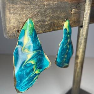 Peut inclure: Une paire de boucles d'oreilles abstraites en forme de tourbillon avec un bord doré. Les boucles d'oreilles sont de couleur bleu sarcelle et jaune.