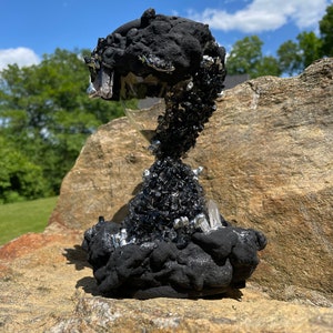 May include: Black hourglass with a crystal-like base and black crystals around the top. The hourglass is sitting on a large rock.