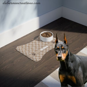 May include: A brown and white dog food mat with a bone and paw print design. The mat has the name "Duke" printed on it. A white ceramic dog bowl with brown dog food is on the mat. A black and brown Doberman Pinscher dog is standing next to the mat.