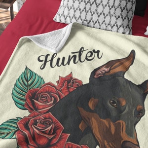 May include: A cream-colored throw blanket with a black Doberman Pinscher illustration. The dog is surrounded by red roses and green leaves. The text "Hunter" is written in black cursive above the dog.