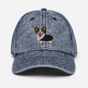 Op de afbeelding: Een baseballcap in denimstijl met een distressed afwerking. De cap heeft een geborduurd beeld van een lachende corgi-hond in zwart, wit en bruin. De cap heeft een gebogen klep en een verstelbare band aan de achterkant.