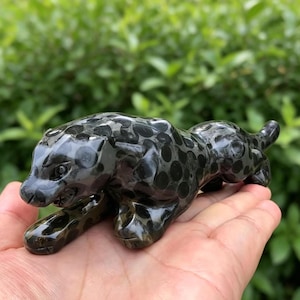 May include: A black and gray spotted jaguar figurine. The sculpture is made of a polished stone material and is in a resting pose. The figurine has detailed facial features and a sleek, smooth finish.