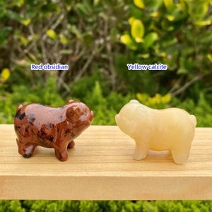 May include: Two small pig figurines. One is red obsidian with black spots, and the other is yellow calcite. The text "Red obsidian" and "Yellow calcite" are above each pig. Both pigs are on a wooden surface.
