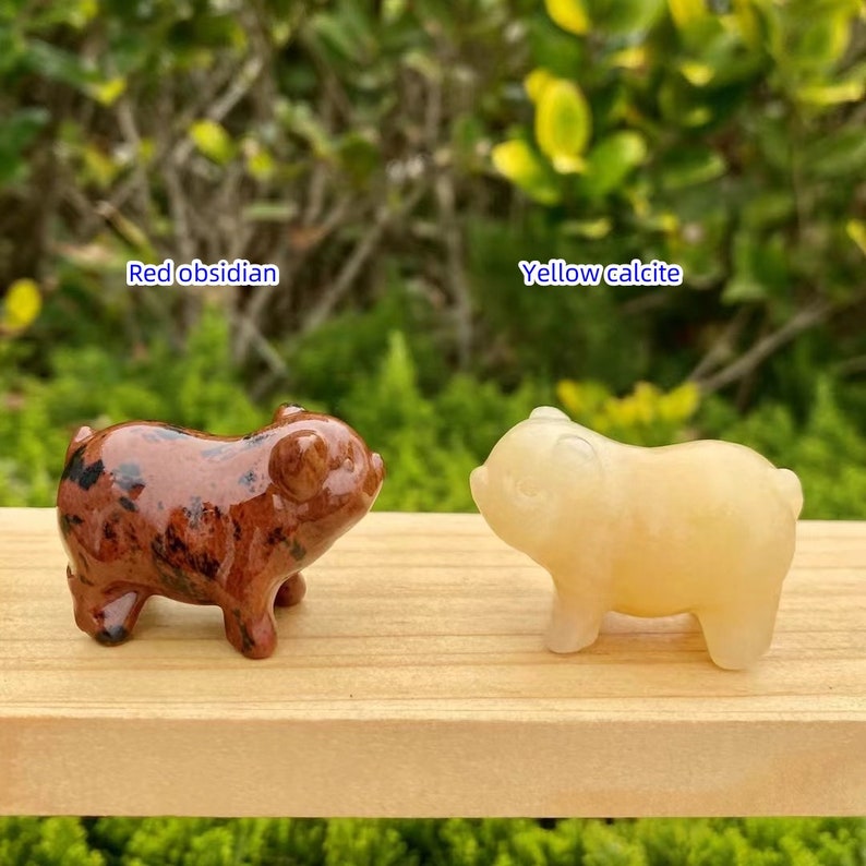 May include: Two small pig figurines. One is red obsidian with black spots, and the other is yellow calcite. The text "Red obsidian" and "Yellow calcite" are above each pig. Both pigs are on a wooden surface.