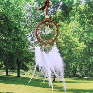 May include: A small brown dreamcatcher with white feathers and a clear crystal hanging in the center. The dreamcatcher is hanging from a brown leather cord and is surrounded by green trees.