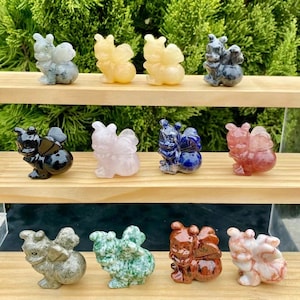 May include: Twelve small, carved crystal bee figurines in various colours, including black, white, yellow, pink, blue, red, green, and grey. The figurines are arranged in three rows of four on a wooden shelf.