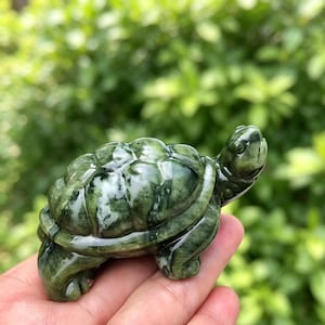 May include: A polished green stone carving of a turtle with a smooth, rounded shell featuring natural white and dark green marbled patterns. The figurine displays a detailed head, four legs, and a glossy finish, showcasing the stone's texture.
