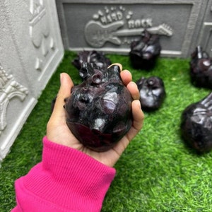 May include: A hand holding a black and red heart-shaped obsidian crystal. The crystal is smooth and polished, with a natural, organic shape. The background is a green grassy surface.