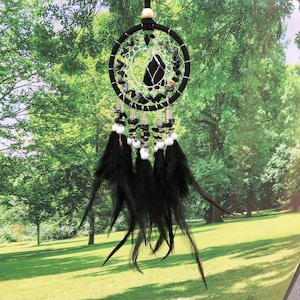 May include: Black dreamcatcher with black feathers and white beads hanging from a black and white beaded hoop. The dreamcatcher is hanging in front of a green grassy background.