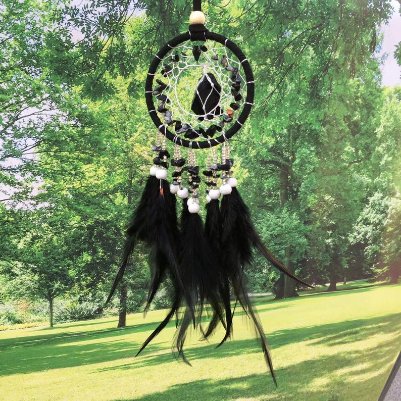 May include: Black dreamcatcher with black feathers and white beads hanging from a black and white beaded hoop. The dreamcatcher is hanging in front of a green grassy background.
