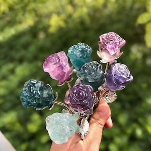 以下が含まれることがあります： 紫、青、緑の色合いの彫刻されたクリスタルローズのブーケ。バラは銀色の茎と葉でまとめられています。背景はぼやけた緑色です。