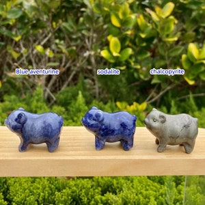 May include: Three small pig figurines made of blue aventurine, sodalite, and chalcopyrite gemstones. The blue aventurine and sodalite pigs are a deep blue color, while the chalcopyrite pig is a metallic gray. The figurines are displayed on a wooden surface.