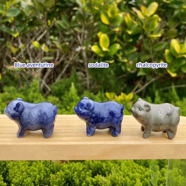 May include: Three small pig figurines made of blue aventurine, sodalite, and chalcopyrite gemstones. The blue aventurine and sodalite pigs are a deep blue color, while the chalcopyrite pig is a metallic gray. The figurines are displayed on a wooden surface.