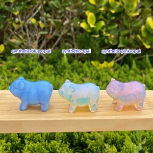 May include: Three synthetic opal pig figurines in blue, iridescent, and pink hues. The figurines are displayed on a wooden surface with text above each pig that reads "synthetic blue opal", "synthetic opal", and "synthetic pink opal".