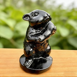 Puede incluir: Una figura negra brillante de un castor, sosteniendo algo en sus patas. El castor está de pie sobre una base redonda. La figura tiene una superficie lisa y reflectante.