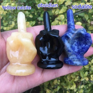 Puede incluir: Tres esculturas de dedo medio talladas en diferentes tipos de piedra. La escultura de la izquierda es de calcita amarilla, la escultura del medio es de obsidiana y la escultura de la derecha es de sodalita.