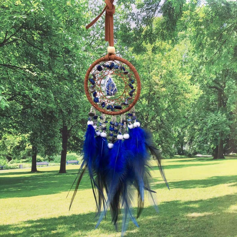May include: A blue and brown dreamcatcher with a lapis lazuli stone in the center. The dreamcatcher is hanging from a brown leather cord and has blue feathers.