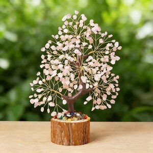 May include: A decorative tree sculpture with a brown trunk and branches made of gold-colored wire. The tree's leaves are small, light pink stones. The tree sits on a wooden base with colorful stones at the base of the trunk.