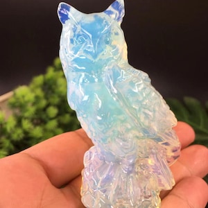 Könnte beinhalten: Eine transluzente, irisierende Eulenfigur. Die Eule ist aus hellblauem und weißem Material geschnitzt, mit Andeutungen von Gelb und Lila. Die Eule sitzt auf einem kleinen Sockel, mit detaillierten Feder- und Gesichtsmerkmalen. Die Figur wird in einer Hand gehalten.