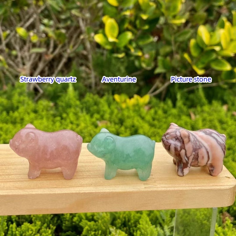 May include: Three small pig figurines made of different stones: strawberry quartz, aventurine, and picture stone. The strawberry quartz pig is pink, the aventurine pig is green, and the picture stone pig is a mix of pink and brown.